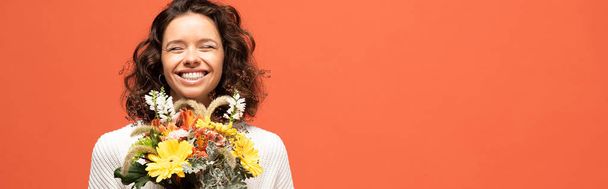 panoramic shot of happy woman in autumnal outfit holding bouquet of flowers isolated on orange - Photo, Image