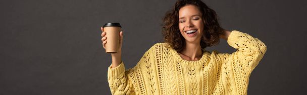 panoramic shot of happy curly woman in yellow knitted sweater with closed eyes holding coffee to go on black background - Photo, Image