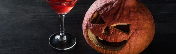 Panoramaaufnahme von geschnitztem Halloween-Kürbis und rotem Cocktail auf schwarzem Hintergrund - Foto, Bild