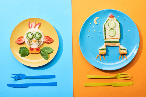 vista superior de platos con cara de lujo y cohete hecho de comida para niños desayuno cerca de cubiertos sobre fondo azul y naranja
 - Foto, Imagen