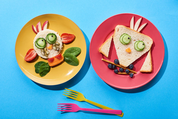 vista superior de platos con vaca y pájaro de lujo hechos de comida para niños desayuno cerca de horquillas sobre fondo azul
 - Foto, Imagen