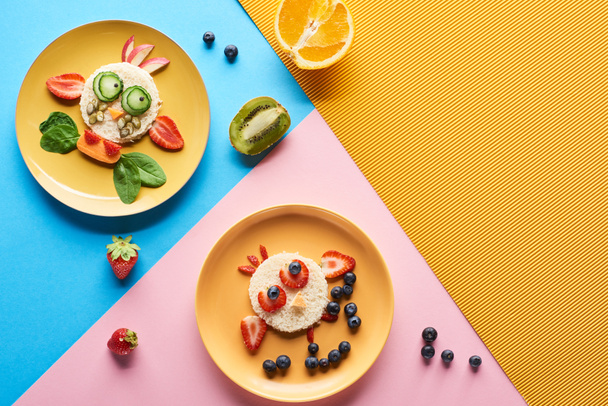 top view of plates with fancy animals made of food for childrens breakfast on blue, yellow and pink background - Fotó, kép