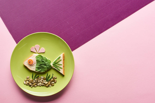 ピンクと紫の背景に食べ物で作られた派手な魚とプレートのトップビュー - 写真・画像