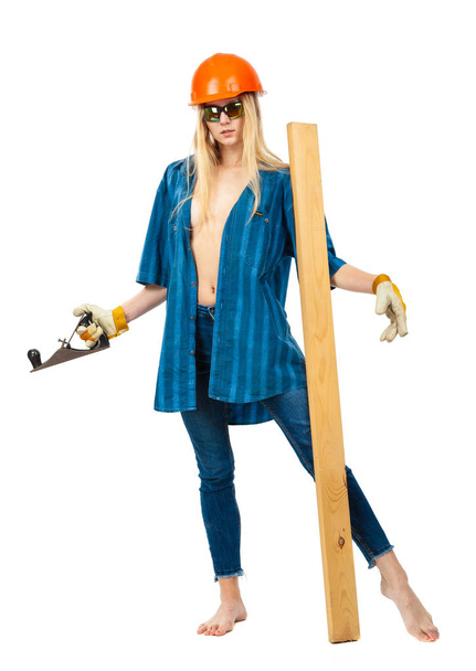 A girl in a construction helmet, with a board and a planer in a blue men's shirt and jeans on a white background. - Valokuva, kuva