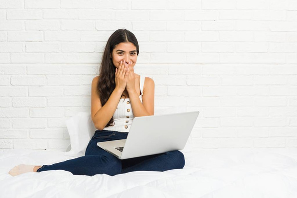 Молода арабська жінка працює зі своїм ноутбуком на ліжку сміється над чимось, покриваючи рот руками
. - Фото, зображення