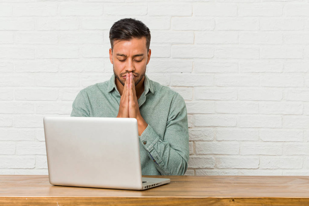Молодий філіппінський чоловік, сидячи на роботі зі своїм ноутбуком, тримає руки в молитві біля рота, відчуває себе впевнено
. - Фото, зображення