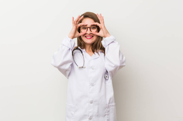 Jonge dokter vrouw tegen een witte muur toont oke teken over de ogen - Foto, afbeelding