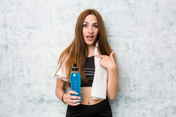 junge kaukasische Sportlerin mit einem isotonischen Getränk überrascht zeigt auf sich selbst, lächelt breit. - Foto, Bild