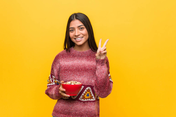 Young arab woman holding a cereal bowl showing victory sign and smiling broadly. - Foto, afbeelding
