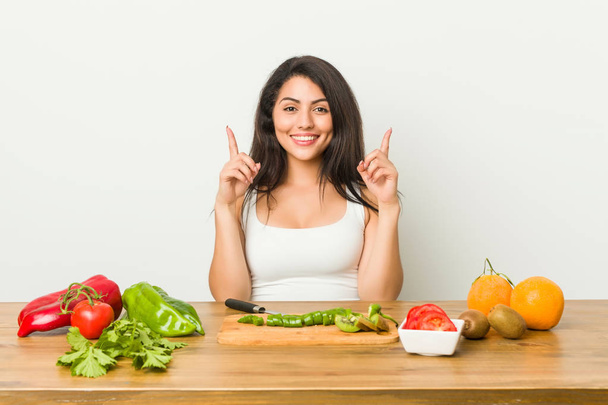 Mujer joven curvilínea preparando una comida saludable indica con los dos dedos delanteros hacia arriba mostrando un espacio en blanco
. - Foto, Imagen