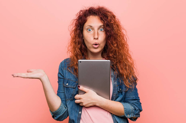 Young caucasian redhead woman holding a tablet impressed holding copy space on palm. - Foto, Bild