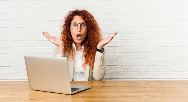 Nuori punapää kihara nainen työskentelee hänen laptop yllättynyt ja järkyttynyt
. - Valokuva, kuva