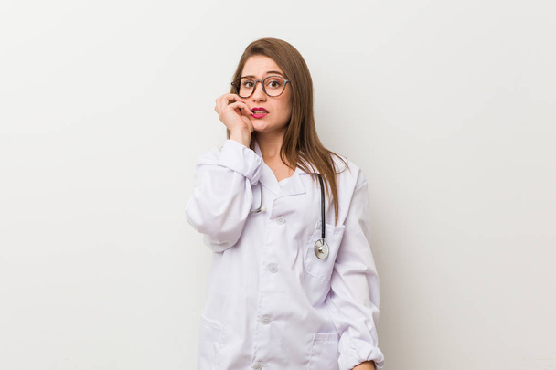 Молодая женщина-врач у белой стены грызет ногти, нервничает и очень беспокоится
. - Фото, изображение