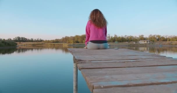 木の桟橋に座っている女性の肖像画は、湖を見て自由に感じます。コンセプトの自由、美しい湖の眺め、幸せな人々、休暇. - 映像、動画