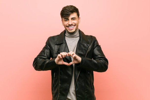Young hispanic man wearing a leather jacket smiling and showing a heart shape with hands. - Foto, imagen
