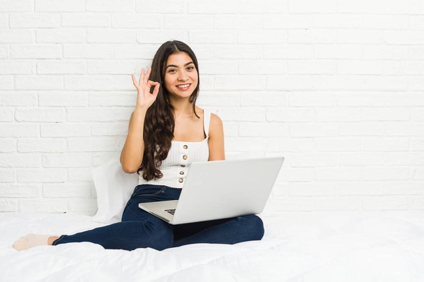 Mujer árabe joven que trabaja con su ordenador portátil en la cama alegre y confiado mostrando gesto ok
. - Foto, imagen