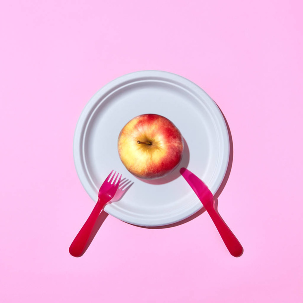 Natural organic apple on a disposable plate served with plastic knife and fork on a pink background with copy space. Top view. - Valokuva, kuva