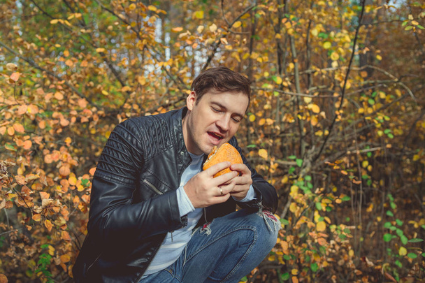 Güneşli bir bahar parkında hamburger yiyen bir adam. Genç erkek, güneşli bir günde sonbahar ormanında hamburger yiyor.. - Fotoğraf, Görsel