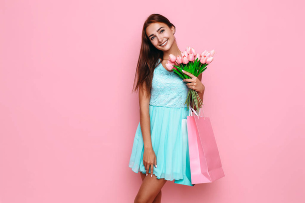 Cute girl in a bright dress holding a bouquet of flowers and shopping bags on a pink background - Fotoğraf, Görsel