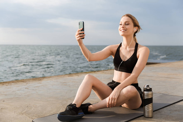 Image of positive woman using earphones and cellphone while sitting on mat near seaside in morning - Foto, imagen