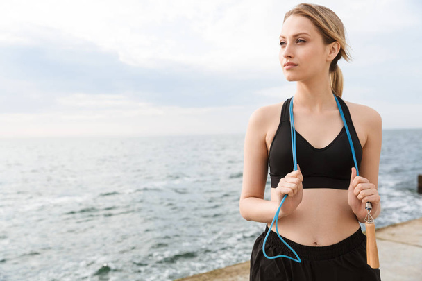 Image of attractive woman standing with jumping rope over neck while working out near seaside in morning - Foto, afbeelding