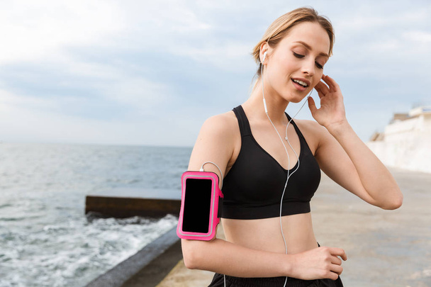 Image of caucasian woman wearing smartphone case listening to music with earphones while jog near seaside - Zdjęcie, obraz