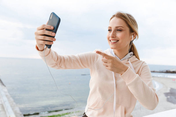 Image of smiling woman listening using earphones and pointing finger at cellphone near seaside in mornin - 写真・画像