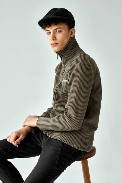 Portrait of handsome teen in black cap and turtleneck posing sitting on chair - Foto, afbeelding