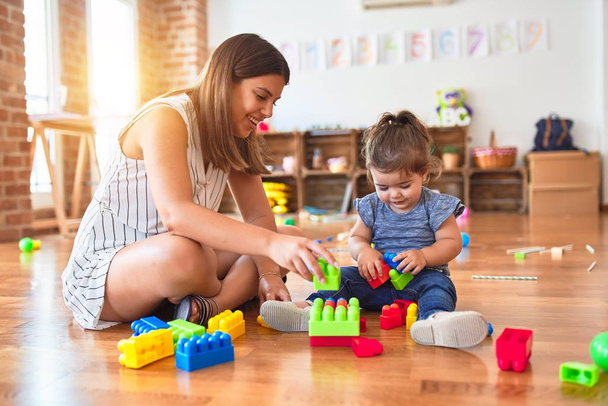 Young beautiful teacher and toddler playing with building blocks toy at kindergarten - Foto, Imagen