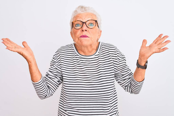Idősebb ősz hajú nő csíkos tengerészeti pólót visel, elszigetelt fehér háttér felett, tanácstalan és zavaros arckifejezéssel, felemelt karokkal és kezekkel. Kétséges fogalom. - Fotó, kép