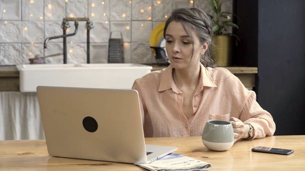 Woman working from home using laptop In the kitchen. Stock footage. Cute brunette woman drinking tea from grey and beige mug, working on laptop, freelance concept. - Photo, Image