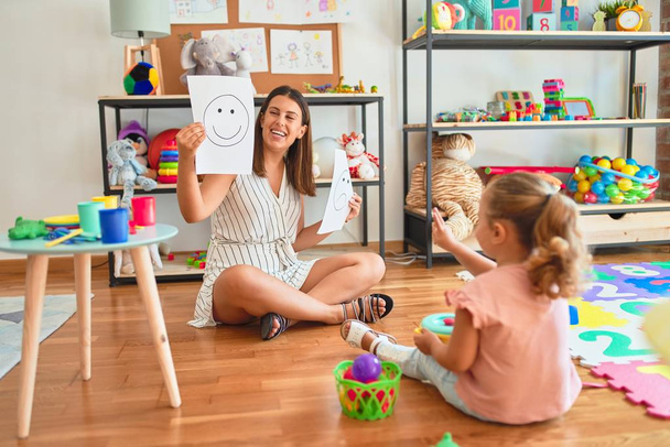 schöne Psychologin und blondes Kleinkind-Mädchen, das auf dem Boden sitzt und im Sprechzimmer eine Therapie mit Emoji-Emotionen macht - Foto, Bild