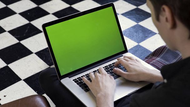 Over the shoulder view of a man using his computer with green screen and sitting on a couch, time lapse. Stock footage. Businessman typing on his laptop with chromakey on checkered floor background. - Photo, Image
