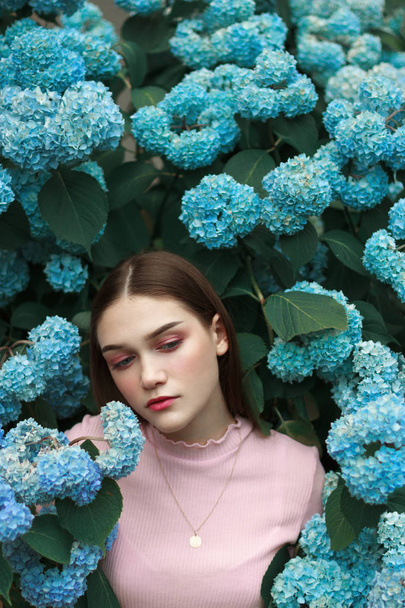 Tendre fille brune de mode avec un maquillage lumineux portant t-shirt rose debout parmi les fleurs bleues avec les yeux fermés
 - Photo, image