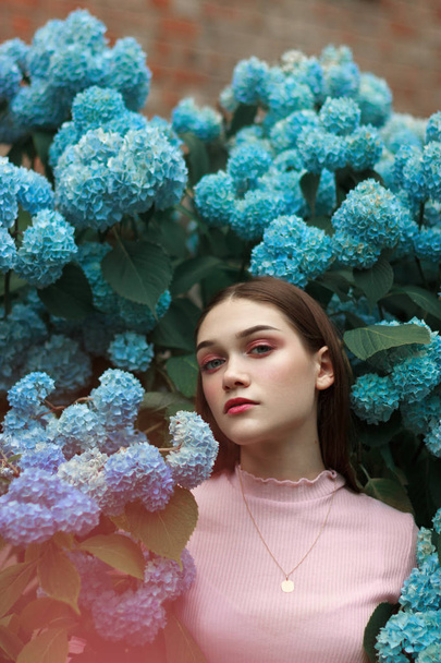 Close up portrait of the beautiful young brunette girl with bright makeup wearing pink t-shirt standing among blue flowers and looking at the camera - Zdjęcie, obraz