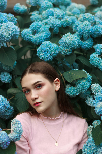 Primer plano retrato de la hermosa joven morena con maquillaje brillante usando camiseta rosa de pie entre flores azules y mirando a la cámara
 - Foto, imagen