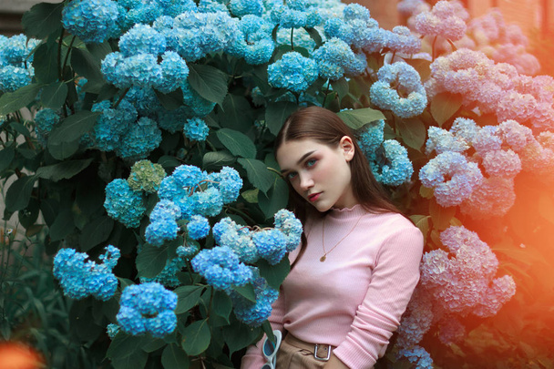 Imagem de menina calma atraente com cabelo morena e maquiagem brilhante vestindo camiseta rosa, de pé entre flores azuis e olhando para a câmera
 - Foto, Imagem