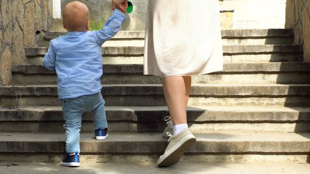 Close up of woman helping her little child boy to climb the stairs by holding his hand. Stock footage. Mother with small kid son walking up the stairs. - Fotó, kép