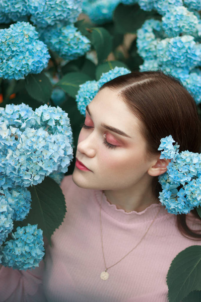 明るいメイクと目を閉じた青い花の間に立つ彼女の耳の後ろに青い花を持つ美しい魅力的な若い女の子 - 写真・画像