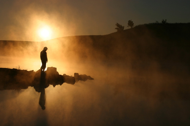 ochtend zon en mist omringen silhouet wandelaar op rustige lake - Foto, afbeelding
