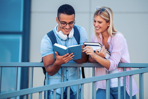 Ευτυχισμένο νεαρό ζευγάρι χαμογελά και διαβάζοντας ένα βιβλίο σε εξωτερικούς χώρους. Απολαμβάνουν την πανεπιστημιακή ζωή. - Φωτογραφία, εικόνα