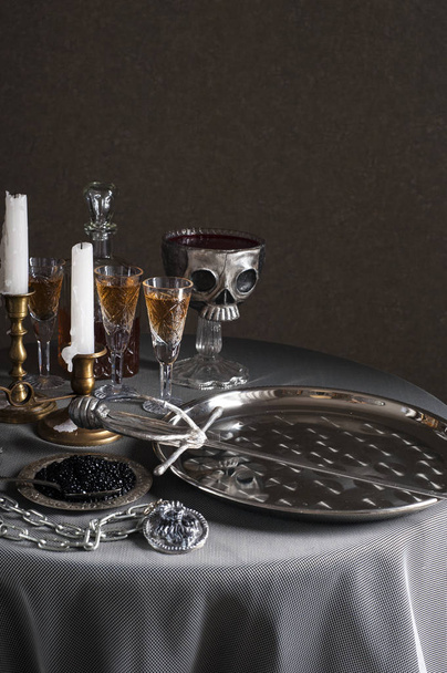 A tray with a sword stands on a round table covered with a tablecloth. Nearby is a silver plate with black caviar and a large medallion with a chain. Behind are candle holders, wine glasses and a skul - Foto, immagini