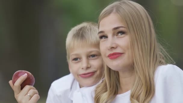 Attractive blond woman sitting with little boy, holding apple in hand and talking. Mother and son spending time together outdoors. Motherhood, parenthood, childhood. - Filmmaterial, Video