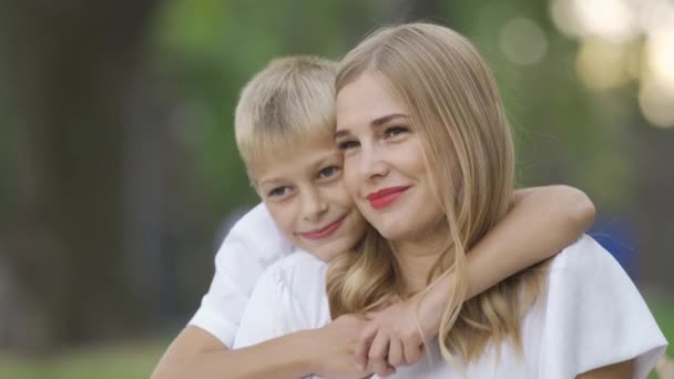 Portrait of young caucasian woman and little boy sitting in the park or forest. Son is hugging his mother. Happy family spending time together. Sunny day outdoors. - Video