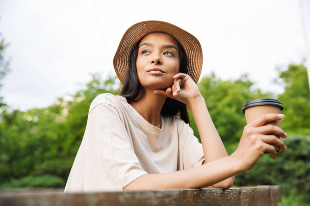 Photo of beautiful woman wearing straw hat holding paper cup with takeaway coffee in green park - Fotoğraf, Görsel