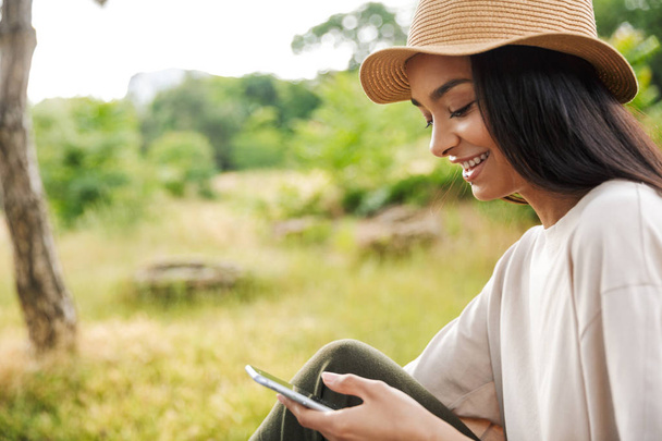 Portrait of charming woman wearing straw hat smiling while holding smartphone in green park - Foto, Imagem