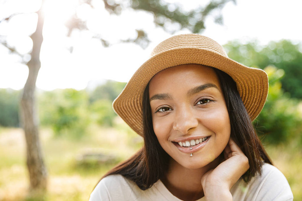緑の公園を歩きながらカメラに微笑む麦わら帽子をかぶった陽気な女性の写真 - 写真・画像