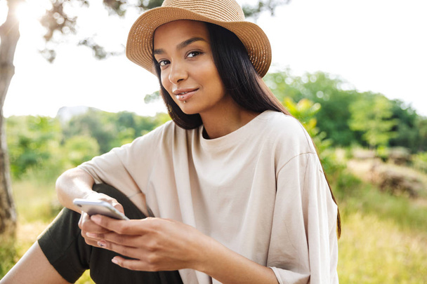 Portrait of pretty woman wearing straw hat holding smartphone while sitting on bench in green park - Φωτογραφία, εικόνα