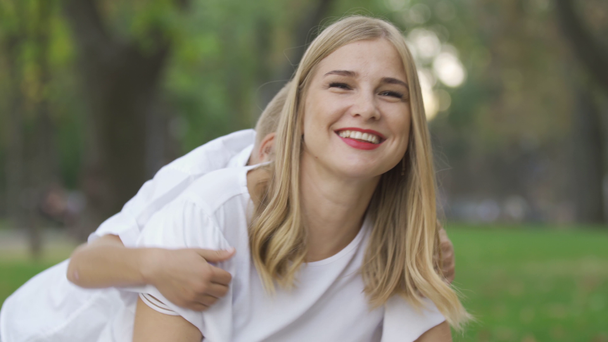 Portrait of attractive caucasian woman in the park looking at camera and smiling. Two little children running towards her from the background and hugging their mother. Sunny day, leisure outdoors. - Video