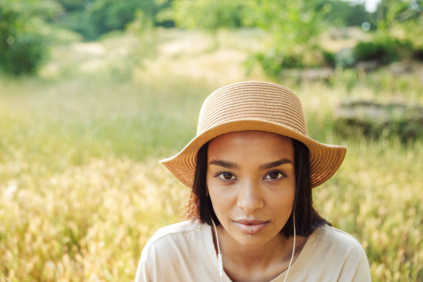 Portrait of beautiful woman wearing straw hat using earphones while sitting on grass in green park - Фото, зображення